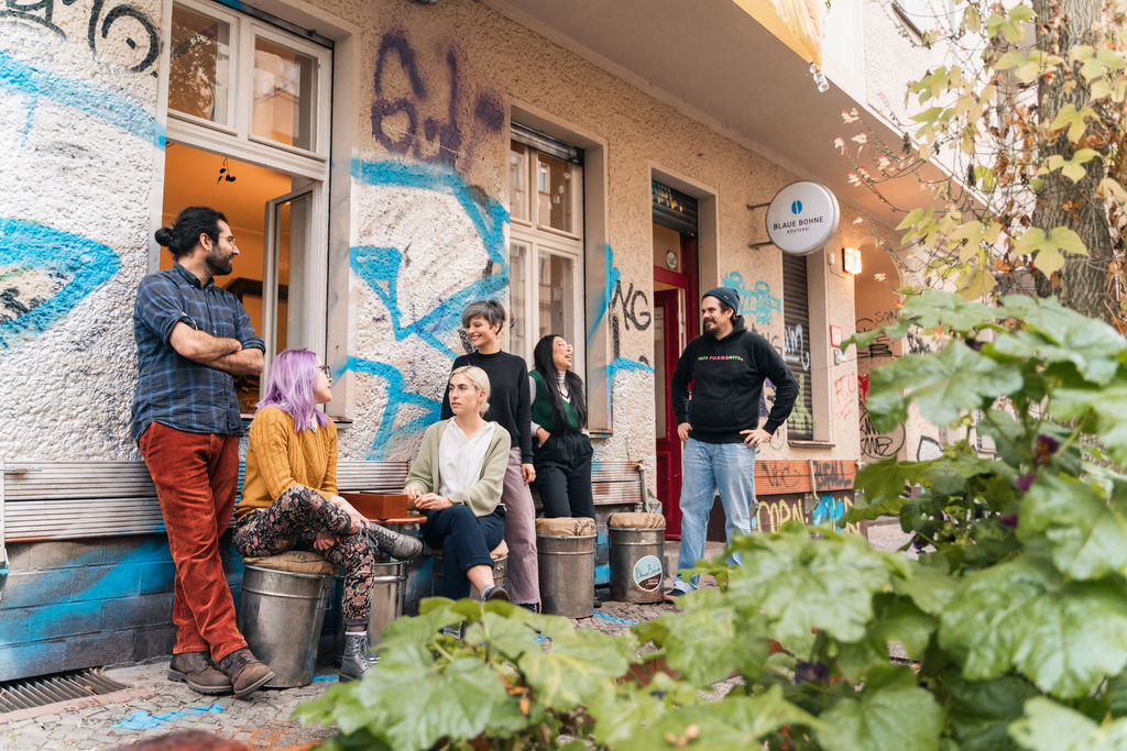 Junge fröhliche Menschen sitzen auf Kaffeebohnen-Tonnen oder stehen vor einem Altbau mit Graffiti und dem Schild „Blaue Bohne Rösterei“ an der Wand und unterhalten sich. Seitlich vorn eine grüne Rankpflanze.