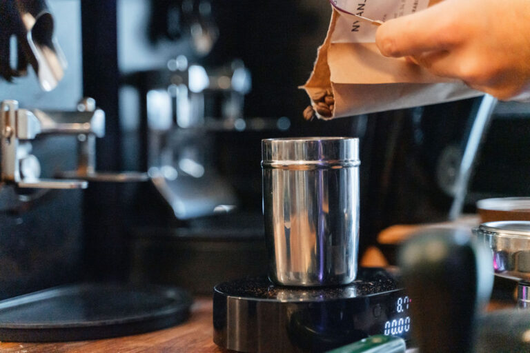Dosierbecher aus Metall auf einer Digitalwaage, in den eine Hand aus einer Papierpackung Kaffeebohnen füllt