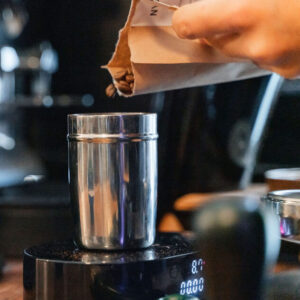 Dosierbecher aus Metall auf einer Digitalwaage, in den eine Hand aus einer Papierpackung Kaffeebohnen füllt