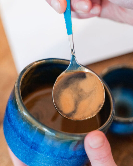 Blaue Tasse mit Espresso, die von einer Hand leicht geneigt wird, während eine andere einen Löffel voll Kaffee mit hellbrauner Crema darüber hält