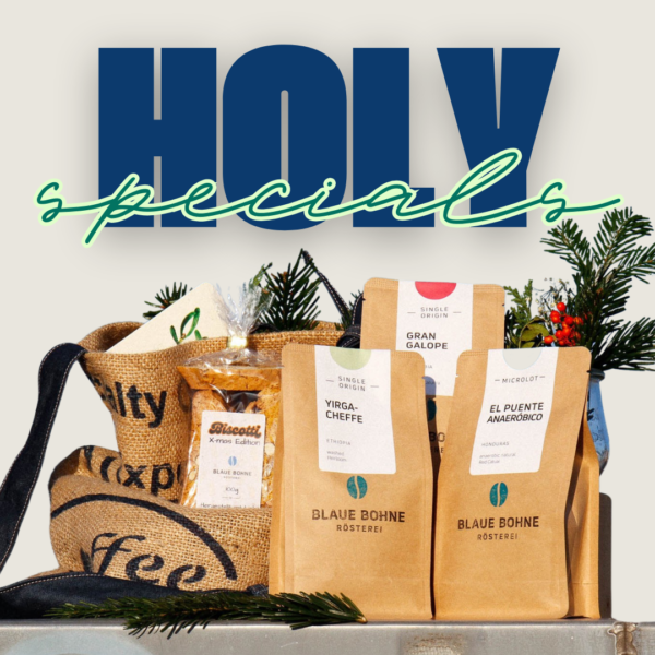 Eine festliche Anordnung des Holy Specials X-mas Coffee Sets von Blaue Bohne mit drei Spezialitätenkaffees: Yirgacheffe, Gran Galope und El Puente Anaeróbico. Dazu Biscotti, eine Weihnachtskarte und eine upgecycelte Kaffeesack-Tasche, präsentiert vor einem strahlenden Himmel und Tannengrün.