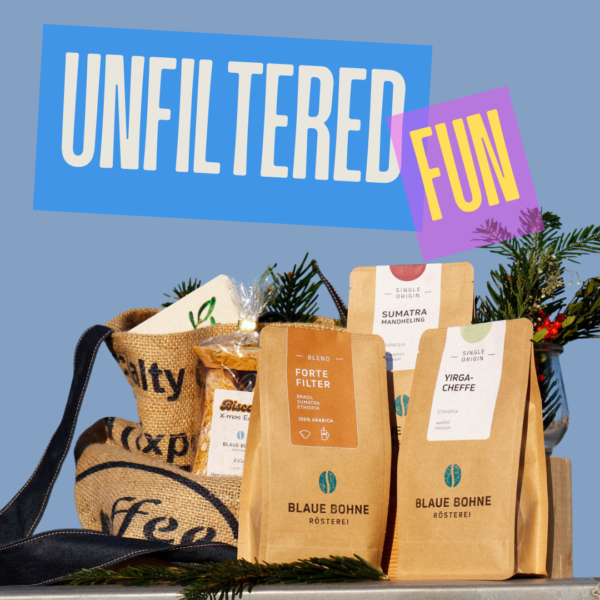 Eine festliche Präsentation des Unfiltered Fun X-mas Coffee Sets von Blaue Bohne, mit Forte Filter, Sumatra Mandheling und Yirgacheffe. Das Set ist stilvoll mit einer upgecycelten Kaffeesack-Tasche, Biscotti, einer Weihnachtskarte und Tannengrün arrangiert.